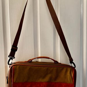 May include: A brown briefcase with a red leather accent and a brown shoulder strap hangs from a hook. The bag has a top handle and a rectangular shape. The edges are trimmed in a lighter brown color.