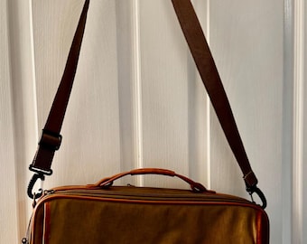 May include: A brown briefcase with a red leather accent and a brown shoulder strap hangs from a hook. The bag has a top handle and a rectangular shape. The edges are trimmed in a lighter brown color.