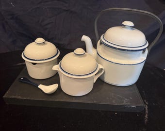 May include: A white enamel tea set featuring a teapot with a metal handle, a lidded sugar bowl, a lidded creamer, and a small spoon. Each piece is accented with a thin navy blue rim detail, providing a clean and uniform appearance for the set.