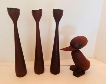 May include: Three tall, dark brown wooden candle holders with a flared top and a small wooden duck sculpture. The duck has a rounded body, a beak, and is perched on a rectangular base. The items are on a white surface.