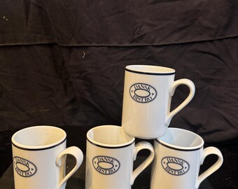 May include: Four white ceramic mugs with a dark blue rim and a central oval logo featuring the text DANSK BISTRO. Each mug has a curved handle and a cylindrical body. The mugs are arranged with three on the base and one stacked on top.