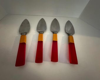 May include: Four cheese knives with stainless steel blades and vibrant handles. The handles transition from a yellow-orange section to a translucent red, creating a striking visual contrast. These knives are designed for serving cheese.