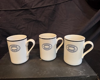 May include: Three white ceramic mugs with a dark blue rim and a central oval logo featuring the text DANSK BISTRO. Each mug has a curved handle on the right side and a smooth, cylindrical body. The design is consistent across all three pieces.