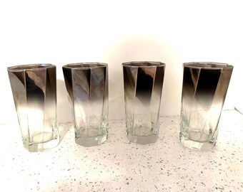 May include: A set of four clear glass tumblers featuring a geometric, faceted design. Each glass has a distinct ombre effect, transitioning from a dark, metallic charcoal gray at the top rim to a completely transparent base at the bottom.