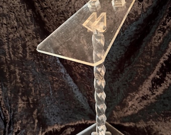 May include: Clear acrylic display stand with a diamond-shaped base and a twisted stem. The top is a clear, angled platform with two small square supports. The stand is designed to hold an item for display.