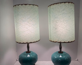 May include: Pair of vintage table lamps. Each lamp features a teal-colored base with floral detailing, a twisted metal stem, and a light blue shade. The shades have a scalloped edge. The lamps sit on a wooden base and have a brown cord.