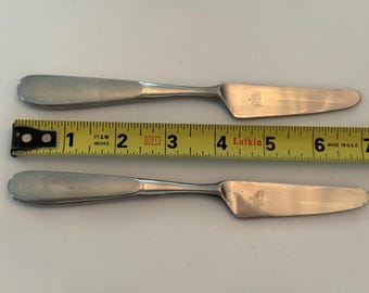 May include: Two silver-toned stainless steel knives with rounded handles and flat blades, each stamped with the Dansk logo. A yellow Lufkin measuring tape is positioned between the knives, showing a total length of approximately 7.5 inches.
