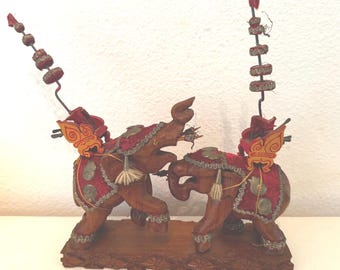 May include: Two wooden elephants facing each other, decorated with red velvet, gold trim, and colorful accents. The elephants are standing on a wooden base.