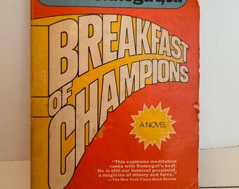 May include: A vintage paperback book titled "Breakfast of Champions" by Kurt Vonnegut, Jr. The cover is orange with yellow and white lettering. A blue banner at the top displays the author's name. A quote from The New York Times is also visible.