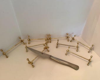 May include: A collection of eight gold-toned metal knife rests, each featuring a central horizontal bar with spherical ends, arranged in a cluster. A single silver-toned stainless steel knife with a pointed blade rests across one of the metal stands.
