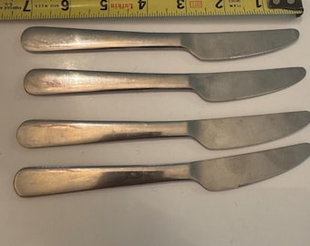 May include: Four silver-toned stainless steel dinner knives arranged horizontally. A yellow Lufkin measuring tape is positioned above the knives, showing they are approximately 7.5 inches in length. Each knife features a smooth handle and a serrated blade.