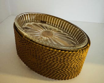 May include: An oval-shaped serving dish with a clear glass insert and a woven gold-colored exterior. The glass insert has a sunburst design in the center. The dish is suitable for serving food or as a decorative piece.