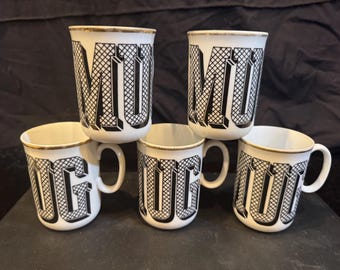 May include: A set of five white ceramic mugs with gold-toned rims. Each mug features a bold, black, three-dimensional graphic pattern forming the letters M, U, and G in a repeating grid design. The mugs are arranged in a stacked formation.