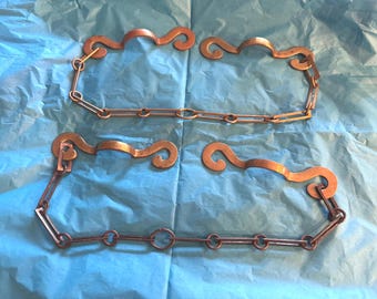 May include: Four copper-colored metal chain links with decorative, curved accents. The links are arranged in two sets of two, each set connected by a chain. The background is a light blue, textured surface.