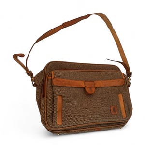 May include: A brown tweed shoulder bag with leather accents. The bag features a long, adjustable leather strap and a flap closure with a buckle. The bag has a rectangular shape and a small logo on the front.
