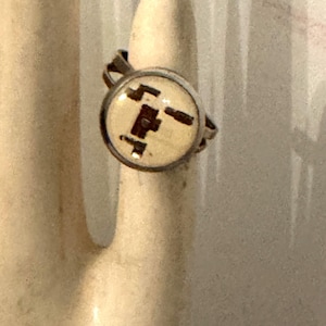 May include: A silver-toned ring featuring a circular setting with a cream-colored inlay. The inlay displays a dark brown, abstract geometric pattern consisting of interconnected rectangular shapes and lines centered within the metal frame.