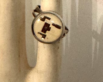 May include: A silver-toned ring featuring a circular setting with a cream-colored inlay. The inlay displays a dark brown, abstract geometric pattern consisting of interconnected rectangular shapes and lines centered within the metal frame.