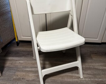 May include: A white folding chair with a slatted seat and backrest. The chair is made of plastic and has a simple, functional design. It is shown in front of a white cabinet.
