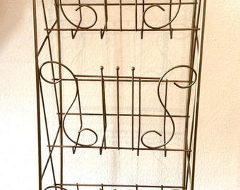 May include: A tall, free-standing metal storage rack finished in a muted gold tone. The unit features three horizontal shelves, each constructed with thin wire bars and decorative scrollwork accents on the sides, supported by four vertical legs.
