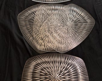 May include: Clear glass serving platters with a radial design. The platters are rectangular with rounded corners. The design radiates from the center of each platter. The background is black.