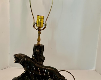 May include: Black ceramic panther lamp with a brass lamp holder. The lamp base features a panther figure on a log with three cup holders. The lamp has a gold-colored metal frame for the lampshade and a brown power cord.