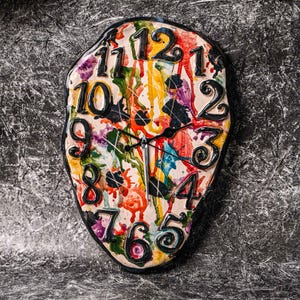 May include: An abstract wall clock with a colourful, splattered paint design. The clock face has black, raised numerals against a white background, with black clock hands. The clock has a black border and a free-form shape.