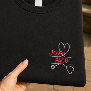 May include: A black crewneck sweatshirt featuring an embroidered design on the lower right side. The design includes a white stethoscope forming a heart shape, with the name Jessica in red script and the text PACU in red block letters below it.