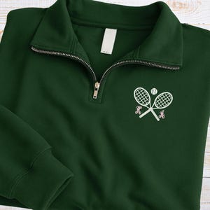 May include: A dark green quarter-zip sweatshirt with a white embroidered tennis racket and ball design. The design includes the letters "K" and "A" in a pink script font. The sweatshirt has a ribbed cuff and a white tag on the collar.
