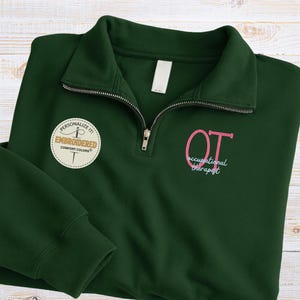 May include: A dark green quarter-zip sweatshirt with the embroidered letters "OT" in pink, and the words "occupational therapist" in a light blue script. The sweatshirt has a white tag and a "Personalize It! Embroidered Comfort Colors" patch.