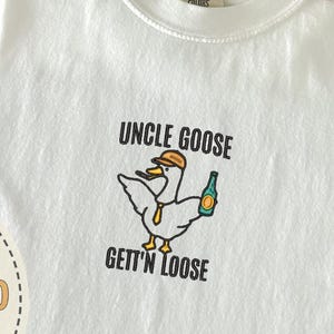 以下が含まれることがあります： 「UNCLE GOOSE GETT'N LOOSE」の刺繍が入った白いTシャツ。帽子をかぶり、ボトルを持った漫画のガチョウが描かれています。Tシャツは、青いジーンズとベージュの帽子の隣に折りたたまれています。