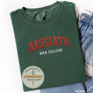 Puede incluir: Una camiseta verde musgo con el texto "BASGIATH WAR COLLEGE" en letras rojas y blancas. La camiseta est&aacute; doblada y colocada junto a un par de pantalones cortos vaqueros azules. La camiseta tiene cuello redondo.