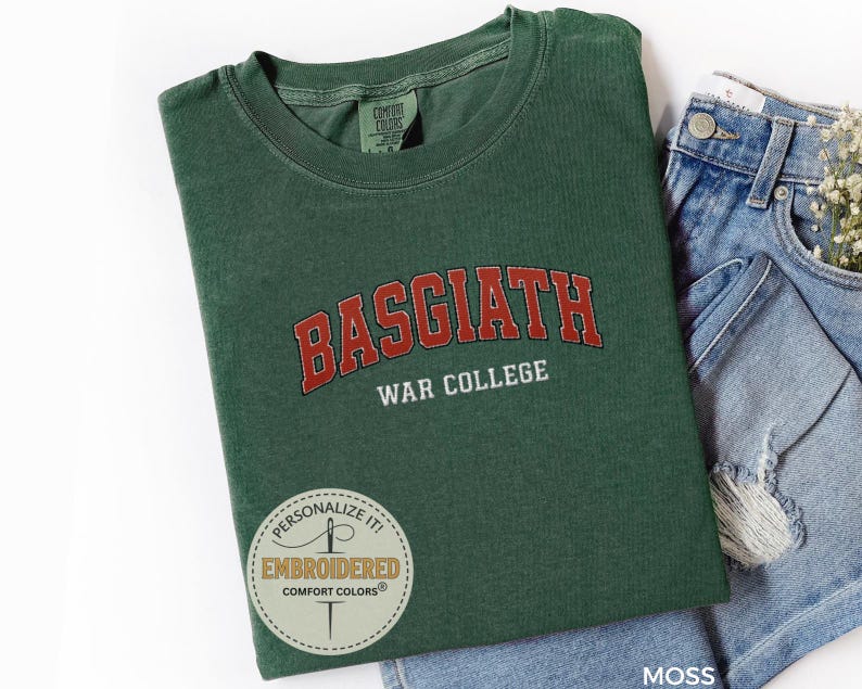 Puede incluir: Una camiseta verde musgo con el texto "BASGIATH WAR COLLEGE" en letras rojas y blancas. La camiseta est&aacute; doblada y colocada junto a un par de pantalones cortos vaqueros azules. La camiseta tiene cuello redondo.