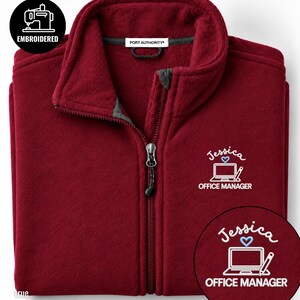 May include: A deep red fleece jacket with a full-length front zipper and a high collar. The chest features an embroidered design with the name Jessica, a light blue heart, a laptop icon, a pencil, and the text OFFICE MANAGER.
