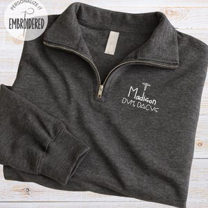 May include: A dark gray embroidered quarter-zip pullover with the text "Madison DVM, DACVS" in white. The pullover has a silver zipper and a small white tag on the collar. The upper left corner has a white circle with the text "Personalize it Embroidered".