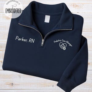 May include: Navy blue embroidered quarter-zip sweatshirt. The text "Parker, RN" is embroidered on the left chest. The right chest features a graphic and the words "Pediatric Case Manager". The top left corner has the text "Personalize it EMBROIDERED".