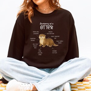 Puede incluir: Una sudadera negra con el texto "Anatom&iacute;a de una nutria" y una ilustraci&oacute;n de una nutria con partes del cuerpo etiquetadas. Las etiquetas incluyen "Generador de sonrisa permanente", "Orbes de detecci&oacute;n de travesuras" y "Aletas de lecho de r&iacute;o".
