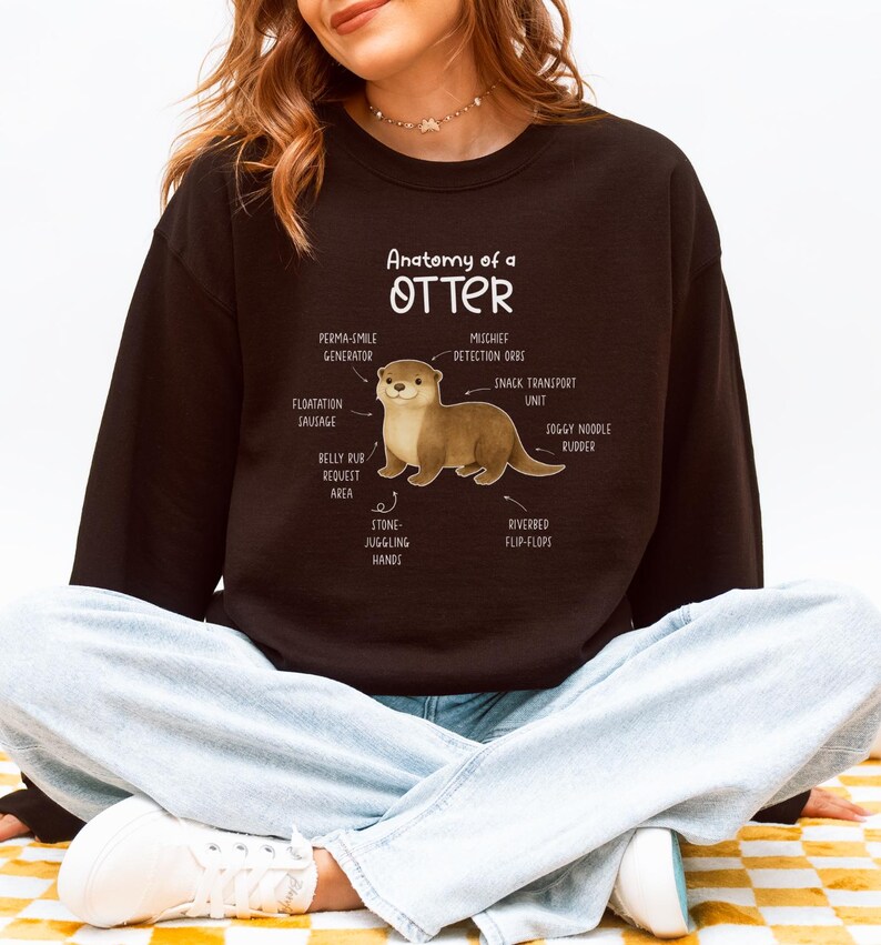 Puede incluir: Una sudadera negra con el texto "Anatom&iacute;a de una nutria" y una ilustraci&oacute;n de una nutria con partes del cuerpo etiquetadas. Las etiquetas incluyen "Generador de sonrisa permanente", "Orbes de detecci&oacute;n de travesuras" y "Aletas de lecho de r&iacute;o".