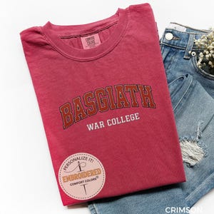 Puede incluir: Camiseta de color carmes&iacute; con el texto "BASGIATH WAR COLLEGE" en una fuente universitaria. La camiseta est&aacute; doblada junto a un par de vaqueros azules con un peque&ntilde;o ramo de flores blancas en el bolsillo. Una pegatina circular dice "PERSONALIZE IT! EMBROIDERED COMFORT COLORS".