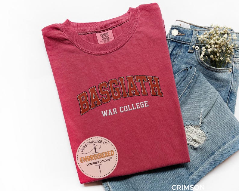 Puede incluir: Camiseta de color carmes&iacute; con el texto "BASGIATH WAR COLLEGE" en una fuente universitaria. La camiseta est&aacute; doblada junto a un par de vaqueros azules con un peque&ntilde;o ramo de flores blancas en el bolsillo. Una pegatina circular dice "PERSONALIZE IT! EMBROIDERED COMFORT COLORS".