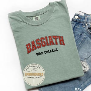 Puede incluir: Camiseta verde salvia con la palabra "BASGIATH" en letras rojas arqueadas sobre "WAR COLLEGE" en negro. La camiseta est&aacute; doblada junto a unos pantalones cortos vaqueros. Una pegatina redonda con texto es visible en la camiseta.