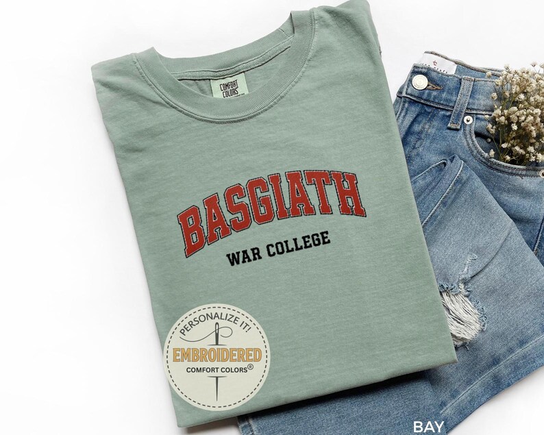 Puede incluir: Camiseta verde salvia con la palabra "BASGIATH" en letras rojas arqueadas sobre "WAR COLLEGE" en negro. La camiseta est&aacute; doblada junto a unos pantalones cortos vaqueros. Una pegatina redonda con texto es visible en la camiseta.