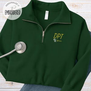 May include: A dark green quarter-zip sweatshirt with the embroidered text "DPT Grace" in yellow. The sweatshirt has a silver zipper and a white tag. A stethoscope is placed on the left side of the sweatshirt.