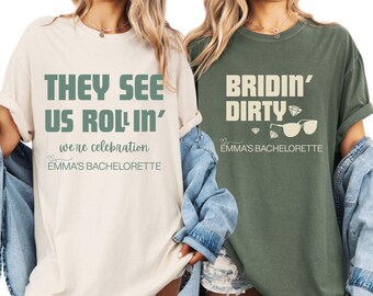 Może przedstawiać: Dwie koszulki, jedna kremowa i jedna oliwkowa, z tekstem na wieczór panieński. Kremowa koszulka mówi "THEY SEE US ROLLIN'" i "we're celebration". Zielona koszulka mówi "BRIDIN' DIRTY" z diamentami i okularami przeciwsłonecznymi. Obie koszulki mówią "EMMA'S BACHELORETTE".