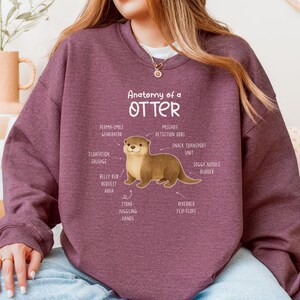 Puede incluir: Una sudadera granate con el texto "Anatomy of an Otter" y una ilustraci&oacute;n de una nutria con partes del cuerpo etiquetadas. La sudadera es de color liso y tiene mangas largas.