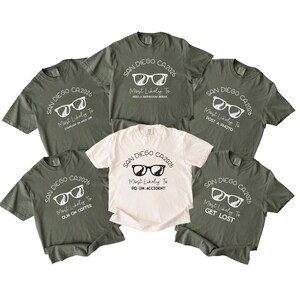 Könnte beinhalten: Eine Sammlung von sechs kurzärmeligen T-Shirts in Olivgrün und Creme. Jedes Shirt zeigt eine Grafik mit einer Sonnenbrille, dem Text SAN DIEGO CA 2026 und individuellen Sprüchen wie Most Likely To Need A Bathroom Break, Post A Photo, Run On Coffee, Get Lost, PR On Accident und Complain On Mile One.