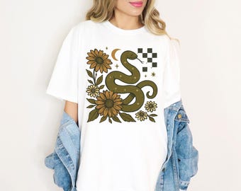 Könnte beinhalten: Weißes T-Shirt mit einem grünen Schlangenmuster, Sonnenblumen, Sternen und einem Mond. Die Schlange ist gewunden und hat eine gespaltene Zunge. Das Design enthält ein kariertes Quadrat und ist auf ein weißes T-Shirt gedruckt.