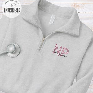May include: A light gray quarter-zip sweatshirt with a white label and the text "EMBROIDERED". The sweatshirt features an embroidered design of "NP" in pink with the name "Emma" below. A stethoscope is visible on the left.