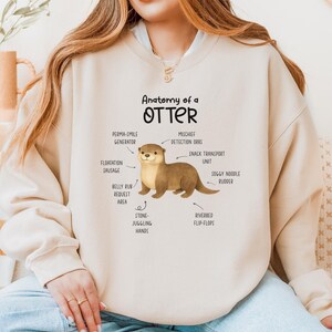 Puede incluir: Sudadera beige con una ilustraci&oacute;n de una nutria y etiquetas anat&oacute;micas humor&iacute;sticas como "Mischief Detection Orbs" y "Soggy Noodle Rudder". El texto "Anatomy of a Otter" est&aacute; impreso encima de la ilustraci&oacute;n. La sudadera tiene mangas largas.
