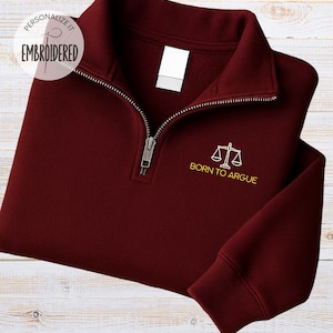Puede incluir: Una sudadera burdeos con cremallera de cuarto de cremallera con una cremallera plateada y las palabras "BORN TO ARGUE" bordadas en dorado, junto con un icono de balanza. La sudadera tiene una etiqueta blanca en blanco.