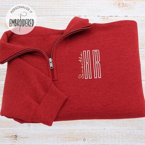 Peut inclure: Un sweat-shirt rouge à fermeture éclair quart de tour avec une broderie blanche sur la poitrine. La broderie comprend le nom Samantha en écriture cursive à côté des lettres capitales HR. Un logo circulaire indique PERSONALIZE IT EMBROIDERED.